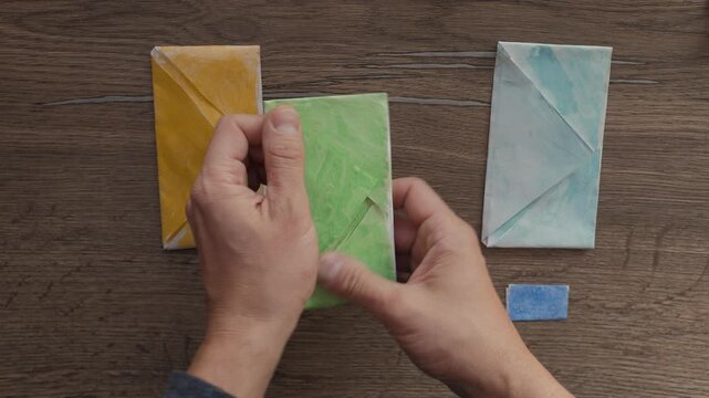 Top-down view of pair of hands organizing three finished origami paper envelopes on textured wooden table, with one hand smoothing green envelope while other steadies yellow envelope in quiet craft