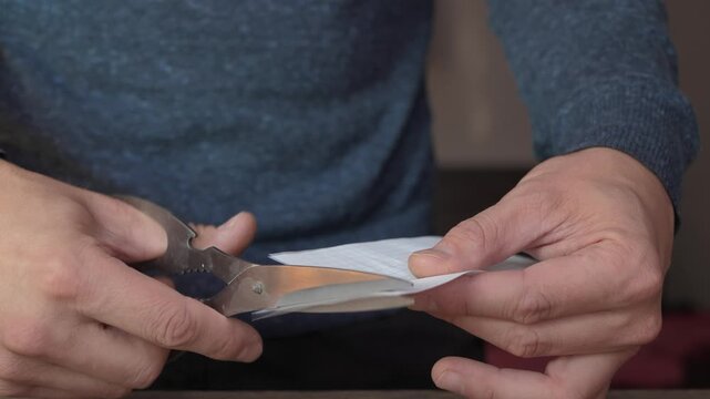 Male hands guiding metal scissors through white paper, blades slicing with steady motion while fingers hold grip, paper bends, separates, and curls during careful craft work