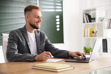 Obraz premium Smiling teacher in suit working with laptop at table in classroom