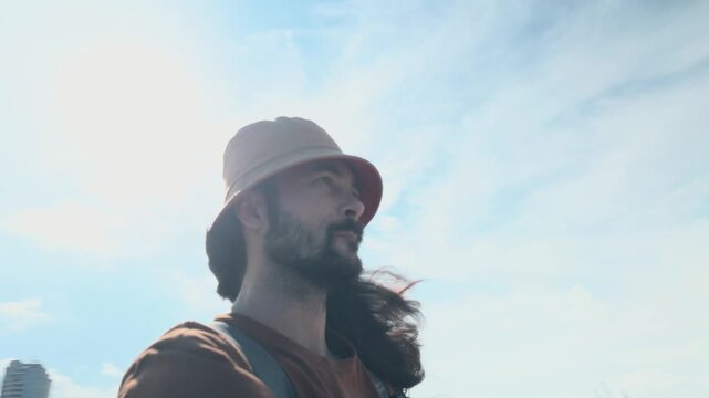 Lone traveler man with long hair and bucket hat walking on harbor pier in coastal city enjoying slow life and freedom near ships cranes and mountains background authentic documentary arc shot