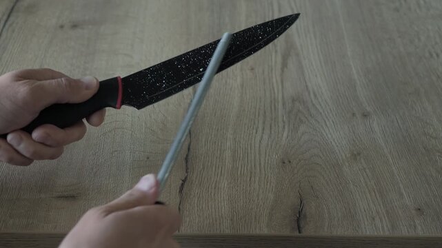 Close overhead view shows two male hands honing kitchen knife on steel rod above wooden cutting board, with repeated controlled strokes, firm grip, careful blade alignment, and clear manual kitchen