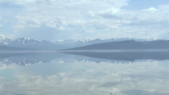 Witness the breathtaking beauty of Khuvsgul Lake, Mongolia. Snowy mountain ranges and a vast cloudy sky are flawlessly reflected on its calm, pristine waters, creating a tranquil tableau of nature.
