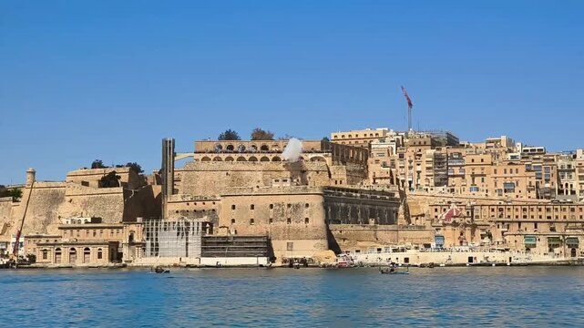 Historic Valletta waterfront with Saluting Battery and Upper Barrakka Gardens, visible cannon smoke above bastions, bright blue sky and harbor setting in Malta