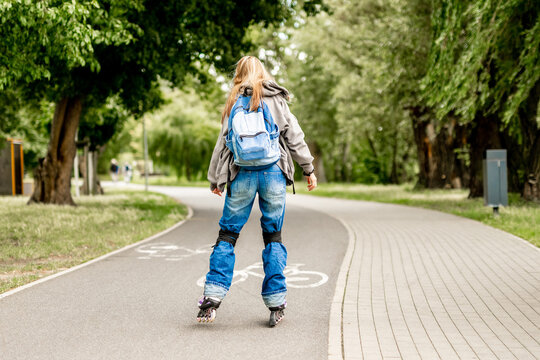 Blonde teen girl rollerblades paved park path. Wears hoodie, jeans, backpack. Green trees visible.
