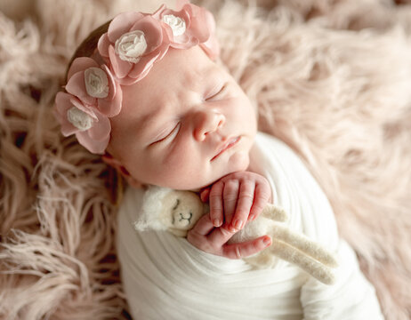 Newborn baby girl wearing flower crown sleeps peacefully holding soft toy on fuzzy pink blanket