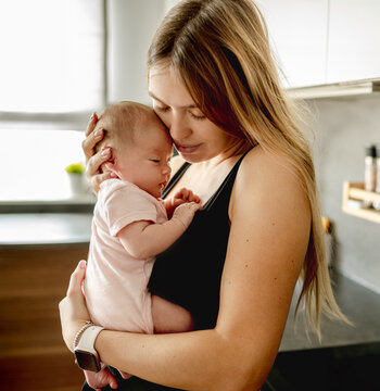 Mother gently cradles her small newborn baby in soft morning light within home kitchen setting