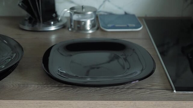 Male hands stack glossy black ceramic plates on a wooden countertop next to a glass cooktop, the shallow glints reflecting the soft, bright kitchen light.
