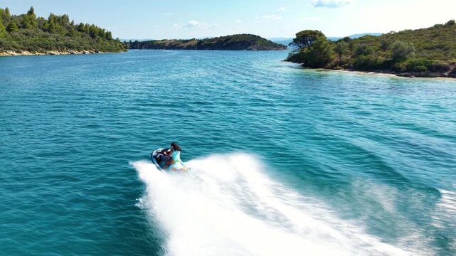 Aerial view of a jet ski speeding across deep blue Mediterranean water, creating a long white wake while riding through a scenic turquoise bay surrounded by green coastal hills.