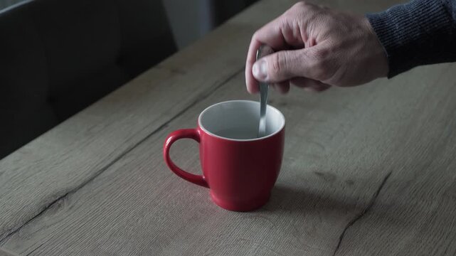 Close view of hand stirring spoon inside red ceramic mug on textured wooden table, showing gentle circular motion, liquid swirl, soft ripples, and warm natural light around cozy drink scene