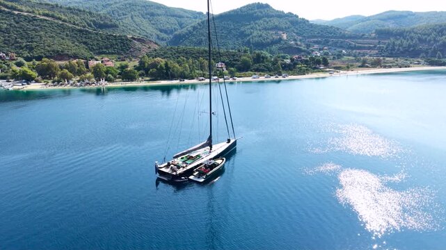 Aerial view of a large sailing yacht anchored with its tender boat in a calm sheltered bay in Sithonia, Halkidiki, Greece, surrounded by clear blue water, sandy beach and forested hills.