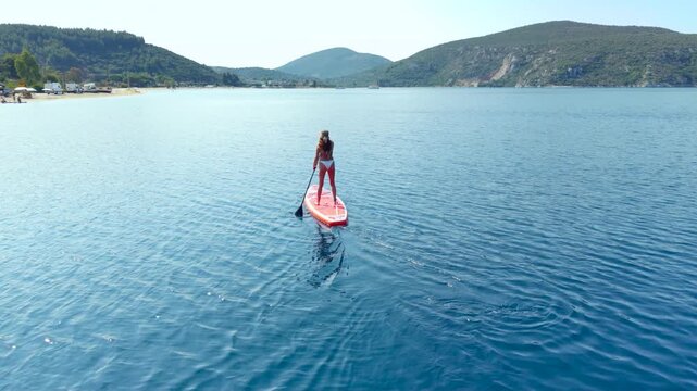 Aerial view of a woman stand-up paddleboarding across calm crystal-clear water in a sheltered bay surrounded by forested mountains in Sithonia, Halkidiki, Greece on a summer day.