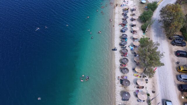 Aerial view of Toroni in Sithonia, Halkidiki, Greece, showing a busy Mediterranean beach with turquoise crystal-clear water, swimmers, sunbeds and beachfront hotels on a sunny summer day.