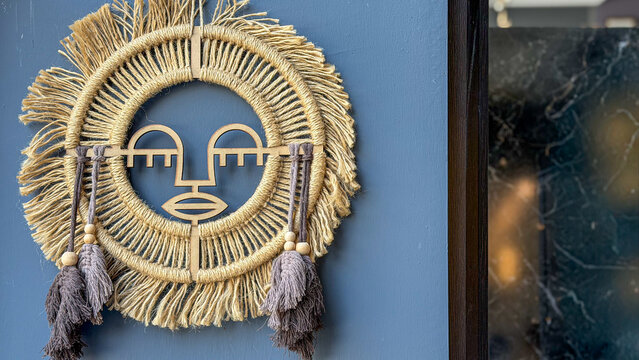 Handcrafted boho wall decoration made of natural rope with an abstract wooden face, tassels and fringe displayed on a blue wall, modern bohemian interior decor.