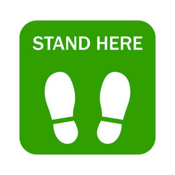 Green square floor sign with white footprints and text that says stand here for social distancing