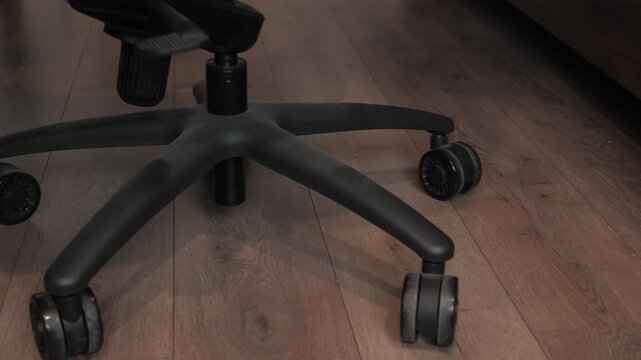 Low-angle close view of office chair five-star base with caster wheels rolling across hardwood floor, showing slow repositioning under desk with soft indoor light, subtle blur on moving wheel