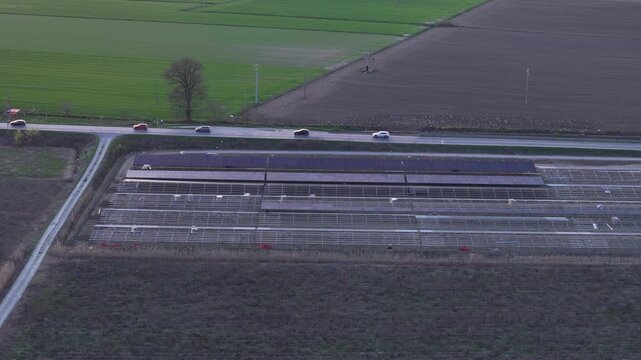 Solar farm construction site shows incomplete photovoltaic mounting rows beside cultivated fields and a rural road, revealing the wider agricultural setting during a super slow motion drone pullback.