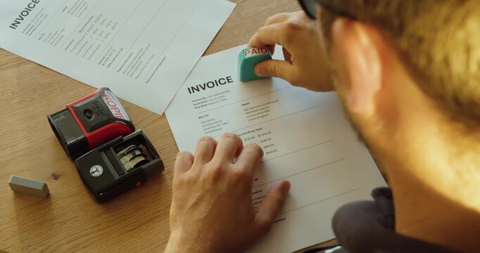 Top view of a man stamping an invoice with a "PAID" stamp after receiving payment. Concept of accounting, bookkeeping, invoice processing, and financial administration.