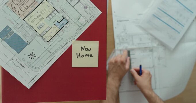 Top-down view of a person reviewing architectural floor plans next to a sticky note labeled "New Home." Concept of home planning, construction, architecture, and property development.