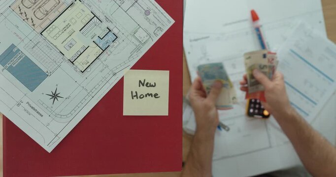 Top-down view of a person calculating finances for a new home while reviewing house plans, counting money, and using a calculator. Concept of home buying, budgeting, and financial planning.