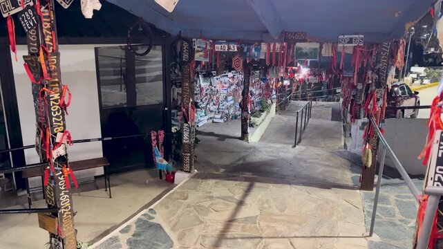 Static right pan capturing illuminated exterior of Difunta Correa Shrine in Vallecito at night, showing hanging prayer bands, votive offerings, and traditional pilgrimage decorations.