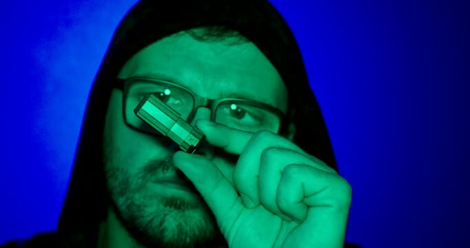 Close-up of a hooded man holding a USB flash drive under neon lighting. Ideal for cybersecurity, hacking, data theft and digital security.