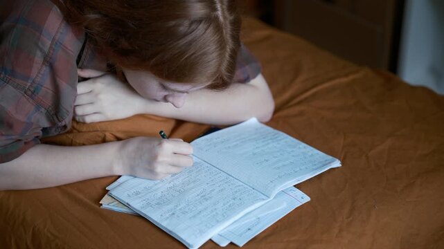 teenage girl studying at home while writing homework in a notebook. The student lies on her bed, solving math exercises and preparing for school in a warm, cozy bedroom. 