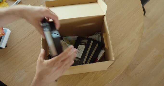 Person opening a cardboard box and looking through old VHS tapes. Ideal for nostalgia, old memories, archives, retro technology and decluttering.