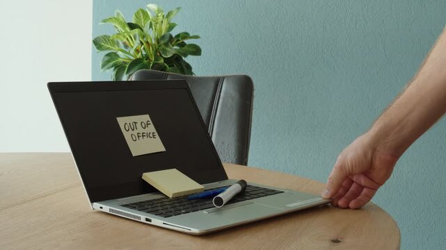 A person places an "Out of Office" sticky note on a laptop before leaving the workspace. Concept of vacation, time off, workplace communication, and temporary absence.