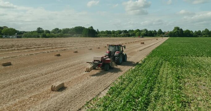 Low-angle drone orbit around a red tractor towing a straw baler across a harvested wheat field under blue skies and cotton clouds in the Dutch countryside on a sunny summer day.