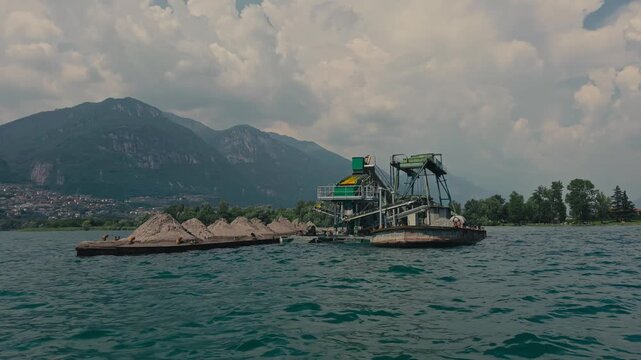 POV from a moving boat passing a working dredger on a tranquil mountain lake, framed by crystal-clear water and spectacular alpine peaks under a sunny summer sky.
