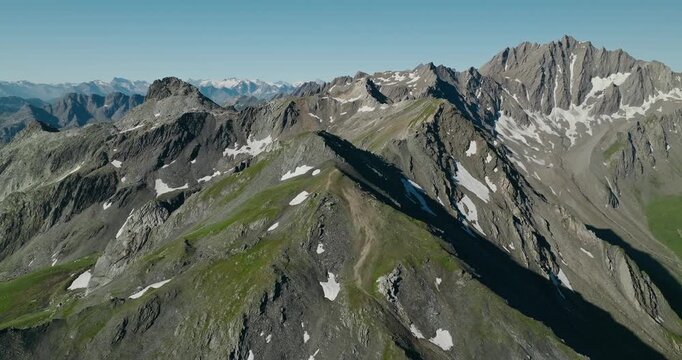 Drone orbits a spectacular Swiss Alpine summit, revealing layered mountain ranges, rugged granite ridges, geological patterns and snowcapped peaks beneath a clear blue summer sky.