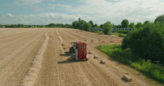 Drone follows a tractor from behind towing a straw baler as rectangular straw bales are ejected onto a harvested wheat field under a clear blue summer sky in the countryside.