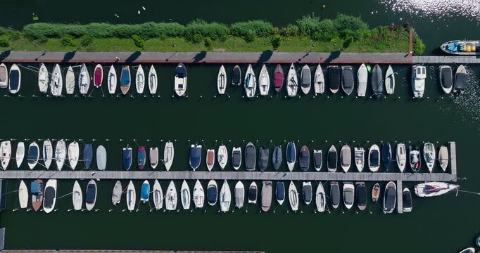 Top-down drone shot showcasing small boats docked in neat parallel rows, creating striking symmetry beside a lakeside parking area with crystal-clear water on a sunny day.