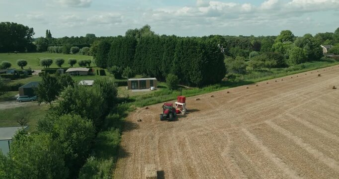 Drone follows a tractor towing a bale accumulator that collects small straw bales and compresses them into larger bales on a harvested wheat field in the Dutch countryside.