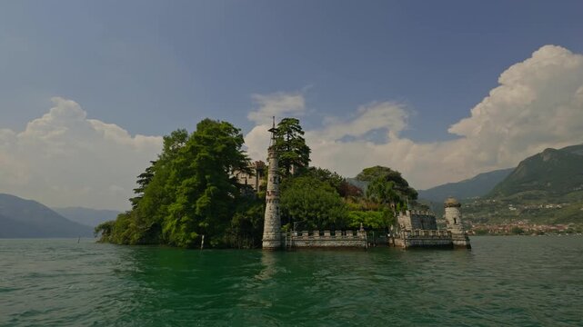 Spectacular view from a boat of the neo-Gothic castle on the tiny Isola di Loreto, framed by mature trees, calm Lake Iseo and majestic alpine mountains beneath a clear blue summer sky.