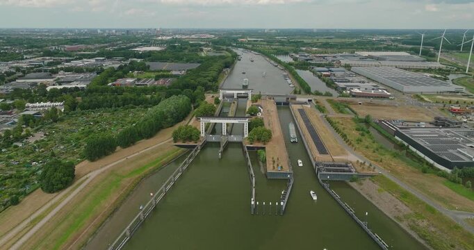 Wide aerial drone view of a cargo ship entering a Dutch sluice with closed gates, leisure boats following behind, surrounded by windmills, industrial estate and rural polder landscape.