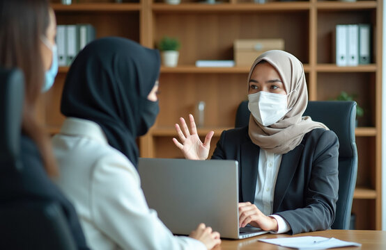 Arab businesswoman in hijab, face mask talks to colleagues during office meeting. Diverse professional team discusses work project at desk with laptop in boardroom. Multiethnic personnel wearing