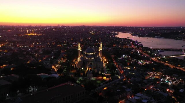 Breathtaking high aerial drone shot of the illuminated Suleymaniye Mosque at night, with glowing city lights stretching toward the Golden Horn waterway during dusk twilight in Istanbul, Turkey.