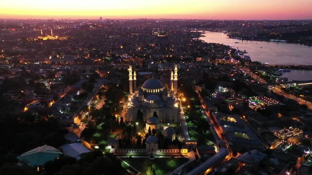 Breathtaking high aerial drone shot of the illuminated Suleymaniye Mosque at night, with glowing city lights stretching toward the Golden Horn waterway during dusk twilight in Istanbul, Turkey.