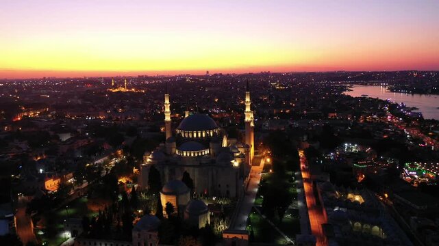Breathtaking high aerial drone shot of the illuminated Suleymaniye Mosque at night, with glowing city lights stretching toward the Golden Horn waterway during dusk twilight in Istanbul, Turkey.