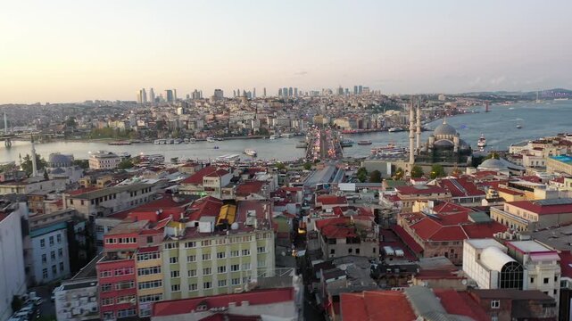 High aerial drone shot over red tile rooftops looking toward Galata Bridge crossing the Golden Horn with Yeni Cami mosque and the Bosphorus strait in Istanbul, Turkey at dusk.
