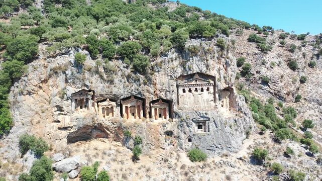 Aerial drone shot of the ancient Lycian rock tombs carved directly into the high cliff faces surrounded by green mountain hillsides in Dalyan, Mugla, Turkey on a sunny day.