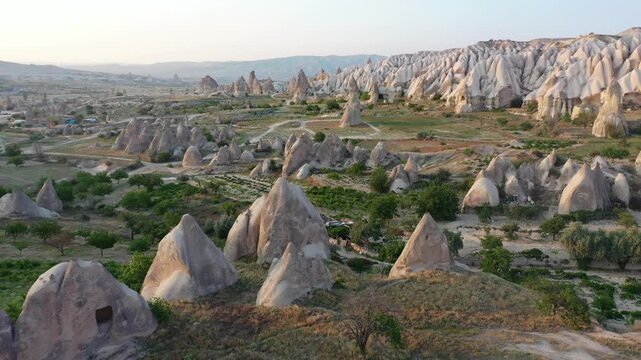 Panoramic aerial drone view of white rock formations, conical fairy chimneys, and flat-topped tuff mountains in Rose Valley, Cappadocia, Nevsehir, Turkey under a clear blue sky.