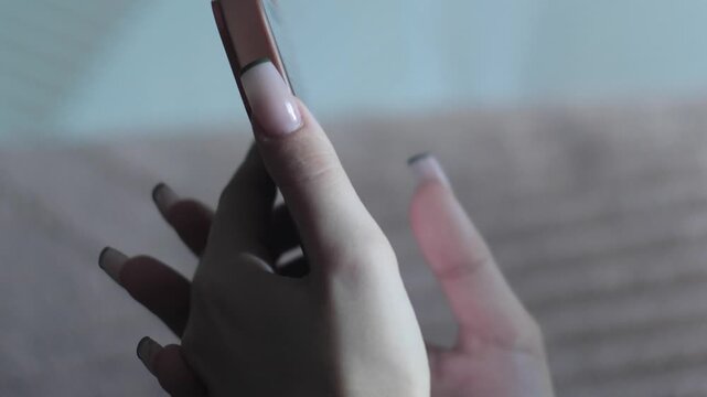 Female hands with an elegant French manicure hold a mobile phone in a brown case. Close-up shot of using a smartphone for reading messages, scrolling, and communicating on social media