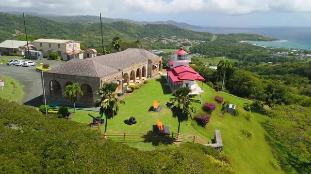 Aerial orbit reveals Fort King George historic lighthouse cannons and barracks overlooking coastline hills of Tobago Caribbean