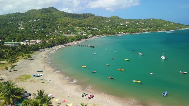 Aerial crane shot descends above Sauteurs Bay revealing turquoise water dotted with colorful fishing boats and yachts along sandy Caribbean beach in Grenada