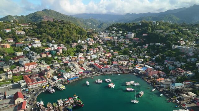 High altitude flyover reveals turquoise Carenage harbor lined with fishing trawlers and yachts framed by colorful hillside St George's town and mountains in Grenada