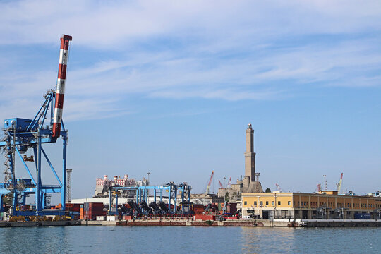 Genoa, Italy - 29 August 2018: View of the historic Lanterna lighthouse overlooking the busy industrial harbor with container cranes, shipping containers, and calm sea.