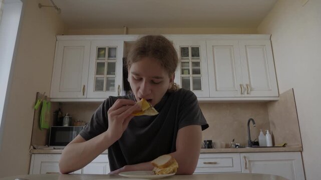 Young person seated at kitchen table eats sandwich, lifts food to mouth, and eats with focused calm in bright home kitchen, hand holding food near chest in soft warm light