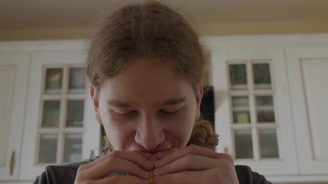 Close-up of young person eating sandwich in domestic kitchen, lowering head after bite with mouth closed, relaxed posture, hands holding food near chest in soft warm light